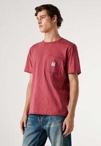 Camiseta roja de manga corta hecha de tejido texturizado con un bolsillo en el pecho que presenta una pequeña etiqueta. Ajuste casual con bordes de dobladillo sin terminar.
