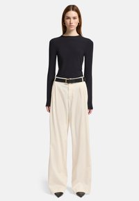 Zwarte blouse met lange mouwen gecombineerd met crèmekleurige broek met wijde pijpen voorzien van plooien en een zwarte riem met een gouden gesp. Puntige zwarte schoenen.