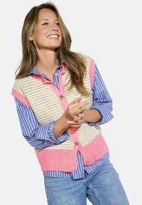 Pletená vesta v krémové barvě s růžovými akcenty a pruhy, s knoflíkovým zapínáním, přes modrou pruhovanou košili. Uvolněný střih s pohodlnou texturou.