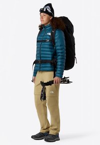 Petroleumkleurige donsjas met horizontale stiksels, beige broek, zwarte rugzak en zwarte wandelschoenen. Houdt trekkingstokken vast; draagt een zwarte muts.