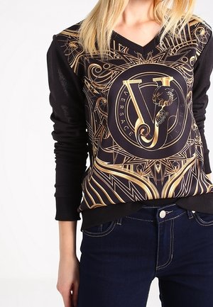 Haut noir à manches longues avec un col en V, orné du logo doré orné de Versace Jeans centré sur la poitrine, associé à un jean bleu foncé.