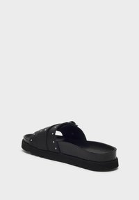 Chancla negra con correas de cuero anchas que presentan tachuelas plateadas y una plantilla texturizada sobre una suela gruesa negra.