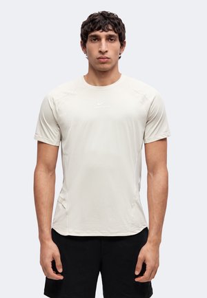 Junger Mann mit kurzen lockigen Haaren, der ein hellbeiges kurzärmeliges Sportshirt und schwarze Shorts trägt und vor einem schlichten Hintergrund steht.