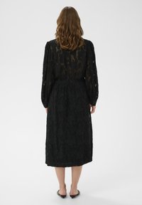 Schwarzes Midikleid mit floralem Muster, langen Ärmeln, elastischem Bund und transparenter Überlagerung. Glatte Textur mit feinen Details. Hausschuh-ähnliches Schuhwerk.