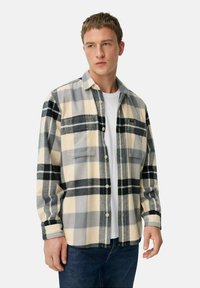 Flanellen shirt in crèmewit, grijs en zwart ruitpatroon; heeft twee borstzakken, knopen en een zachte textuur; gedragen over een wit t-shirt.