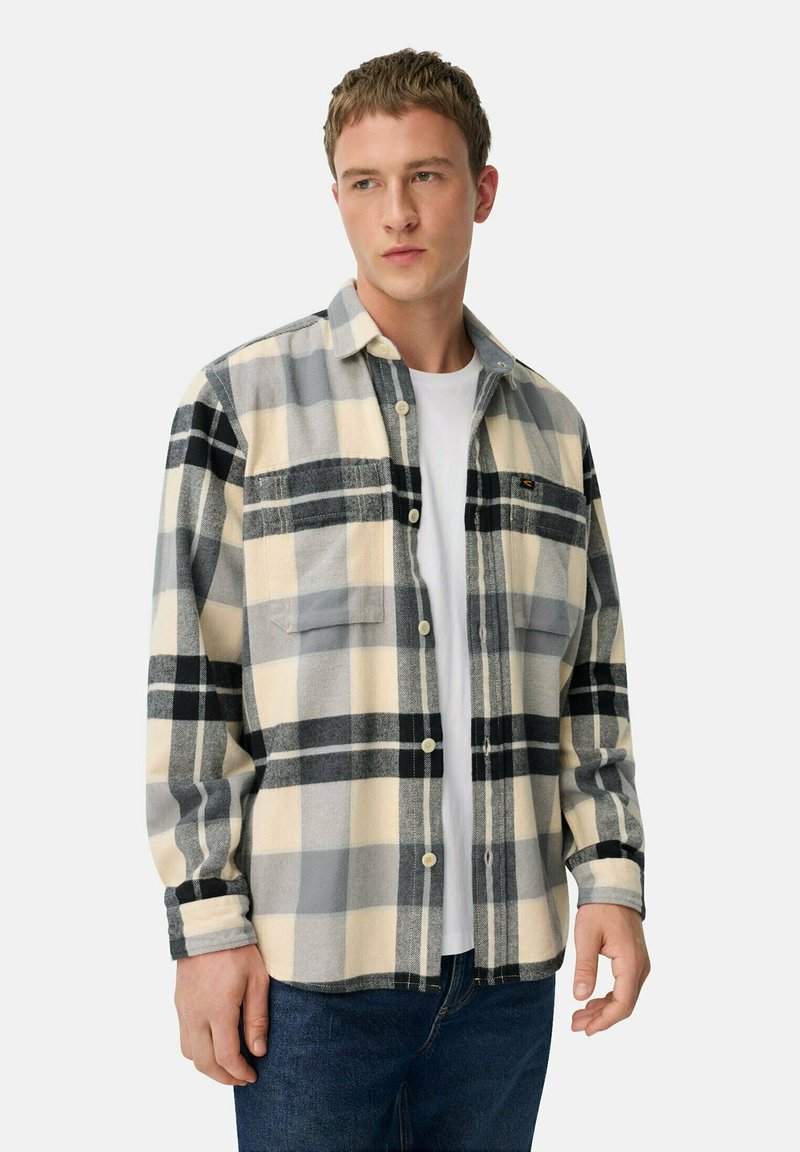 Flanellen shirt in crèmewit, grijs en zwart ruitpatroon; heeft twee borstzakken, knopen en een zachte textuur; gedragen over een wit t-shirt.