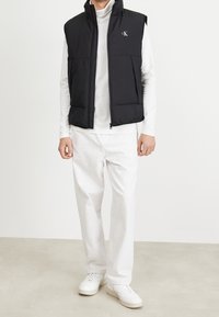 Schwarze gesteppte Weste mit hohem Kragen und Logo, getragen über einem weißen Langarmshirt, kombiniert mit weißen Hosen und weißen Turnschuhen.