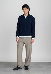 Navy blue pullover with a collar and button placket, paired with light gray, textured trousers and black shoes, set against a neutral background.