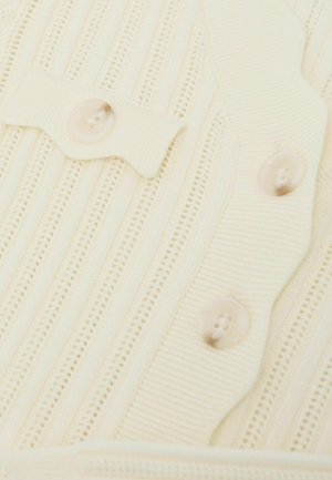 Krémová pletenina s vertikálním žebrováním, vlnitým zdobením a dvěma velkými průhlednými knoflíky na zapínání.