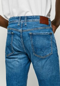 Blugi din denim albastru cu talie înaltă, cinci buzunare și o etichetă din piele pe spate. Prezintă detalii subtile de cusătură și o croială clasică.