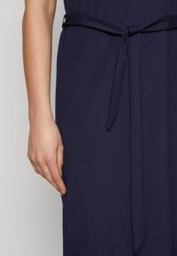 Ναυτικό μπλε φόρεμα με μαλακό, υφασματοποιημένο υλικό, διακοσμημένο με ζώνη στη μέση και εφαρμοστή σιλουέτα, εν μέρει καλυμμένο από ένα χέρι στο πλάι.