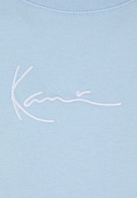 Light blue cotton fabric featuring a white embroidered script logo. The texture is smooth, with a relaxed fit and simple design.