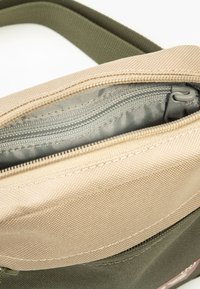 Chaki ir smėlio spalvos audinio krepšys su užtrauktuku, turi lygią vidinę pamušalą, tvirtą siuvimą ir nuimamą diržą.