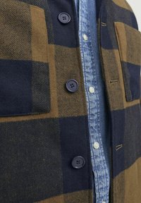 Ruskea ja laivastonsininen ruudullinen takki, joka on valmistettu villasta, sisältäen neljä tummaa nappia, rintataskun ja vaaleansinisen farkkupaidan alla.