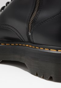 Schwarzlederne Stiefel mit einem seitlichen Reißverschluss, strukturiertem Gummisohle und gelben Nähten. Gebogene Form mit geriffelten Details an der Sohle.