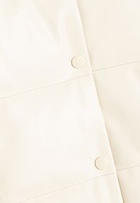 Creme syntetisk læderstof med en glat tekstur, der har synlige sømme og runde trykknapdetaljer.