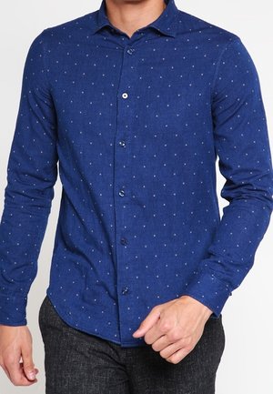 Mężczyzna ubrany w dopasowaną niebieską koszulę na guziki z delikatnym wzorem kropek oraz ciemnoszare teksturowane spodnie, poprawiający rąbek koszuli.