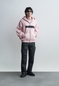 Giovane uomo che indossa un maxi hoodie rosa con zip e la scritta "MISBHV", pantaloni cargo neri, sneakers nere e occhiali da sole rettangolari neri.