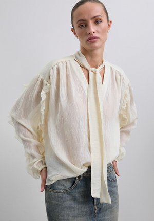 Blusa cremoso y ligera con tela texturizada, mangas acampanadas y un lazo largo en el escote, combinada con jeans de mezclilla azules.