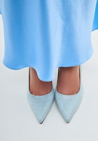 Ljusblå spetsiga klackar dekorerade med små rhinestones, bärs under ett flödande blått tygplagg. Slät textur och elegant design.