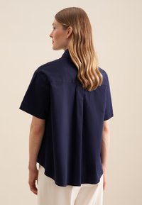 Granatowa koszula z krótkim rękawem, wykonana z lekkiego materiału, z pleceniem z tyłu i zaokrąglonym dołem.