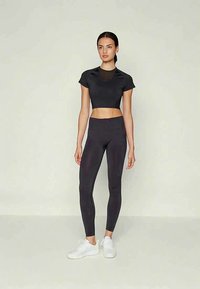 Γυναίκα που στέκεται με μαύρο κοντό αθλητικό τοπ, ταιριαστές κολάν ψηλής μέσης και λευκά αθλητικά παπούτσια σε απλό ανοιχτό φόντο.