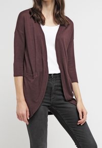 Žena v volném tmavě růžovém kardiganu s tříčtvrtečními rukávy, oblečená přes bílé tričko a černé džíny, stojící na světlém pozadí.