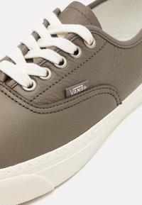 Zapatilla de cuero gris con cordones blancos, que presenta una suela de goma blanca cosida y una pequeña etiqueta con el logo de Vans en el lateral.