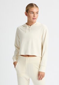 Felpa corta color crema in tessuto morbido con cappuccio regolabile, maniche lunghe e un branding discreto sul petto. Indossata con pantaloni abbinati.