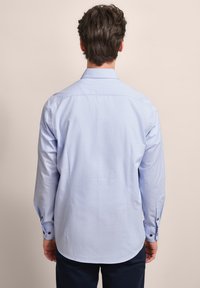 Hellblaues Langarmhemd mit Button-Down-Kragen, abgerundetem Saum und braunen Knopfakzenten an den Manschetten. Glatte Stofftextur.