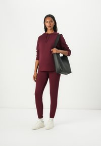 Bordová mikina s kapucňou spojená s vyhovujúcimi nohavicami. Model drží čiernu tašku na rameno. Biele tenisky dopĺňajú outfit.