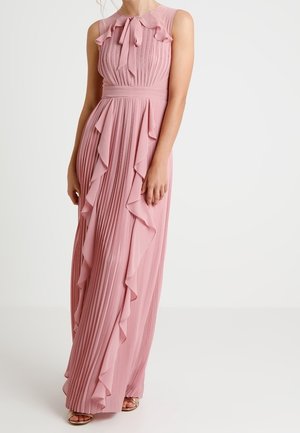 Sleeveless pink maxi dress with pleats, ruffles down sides, and a tied bow at the neckline, worn with open-toe sandals.