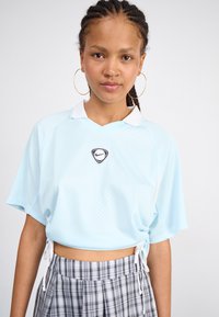Gaiši zils, īss Nike polo ar melnu logotipu, īsām piedurknēm un sānu auklām. Apvienots ar kroku svārkiem šaha rakstā pelēkās krāsās.