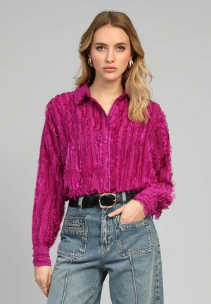 Femme aux longs cheveux blonds porte une chemise boutonnée magenta texturée rentrée dans un jean bleu taille haute avec de grandes poches avant et une ceinture noire.