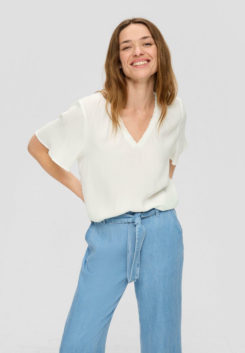 Weißes kurzärmliges Bluse mit V-Ausschnitt und Rüschendetail, kombiniert mit hellblauen, hochtaillierten Hosen mit einem gebundenen Gürtel.