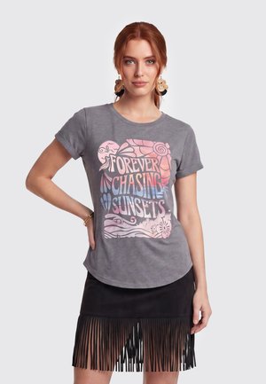 Vrouw met rood haar draagt een grijs T-shirt bedrukt met "Forever Chasing Sunsets" en een zwarte rok met een lange franjeszoom.