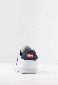 Basket blanche avec un accent au talon bleu marine, tige en textile et synthétique, dotée d'un patch rouge avec un logo de crocodile et des détails texturés.