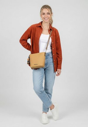 Lächelnde Frau trägt ein rostfarbenes Hemd über einem weißen Oberteil, hellblaue Jeans, weiße Sneakers und eine beige Umhängetasche mit Perlendetail.