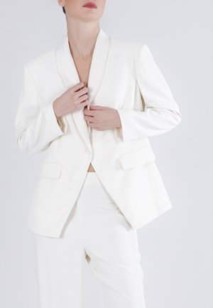 Person iført en hvid skræddersyet dragt med en blazer og bukser, der justerer blazerens krave, mod en enkel lys baggrund.