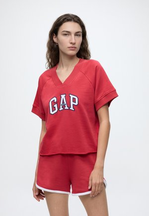 Young woman with long wavy hair wearing red GAP short-sleeve top and matching shorts with white trim against plain background.