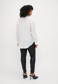 Camicia bianca a maniche lunghe con orlo curvo, tessuto testurizzato e polsini regolabili, abbinata a leggings neri lucidi e sandali con tacco.