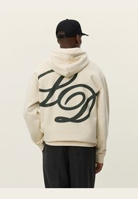 Crème-kleurige hoodie met een groot donkergroen gestileerd logo op de achterkant. Zachte stof met een ontspannen pasvorm en een capuchon met trekkoorden.
