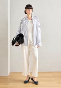 White button-up shirt with blue stripes, cream wide-leg trousers, and dark espadrille-style shoes. Model holds a black shoulder bag.
