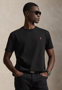 Jeune homme portant des lunettes de soleil noires, un t-shirt noir avec un logo rouge, un pantalon noir et une montre-bracelet, debout les mains dans les poches.