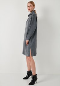 Graues Strickkleid mit langen Ärmeln, einem Kragen, seitlichen Schlitzen und einer lässigen Passform. Getragen mit schwarzen, hohen Stiefeletten.