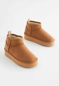 Brune ruskinds støvler til anklerne med en blød pelsforet, rund tå, elastisk bånd og en tekstureret gummisål, der har en dekorativ flap bagpå.