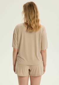 Beżowa bluzka z krótkim rękawem o luźnym kroju, w połączeniu z dopasowanymi szortami. Materiał wydaje się miękki i gładki, idealny na odzież do wypoczynku.