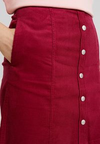 Červená kordurová sukně s přiléhavým střihem, která má pět stříbrných knoflíků vpředu a postranní kapsy. Texturovaná látka s vertikálním žebrováním.