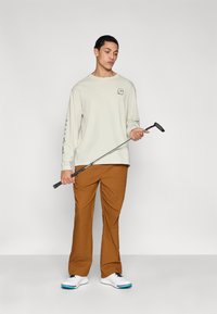 Chemise beige clair à manches longues avec logo, pantalon marron et chaussures blanches avec accents bleus. Le modèle tient un club de golf positionné en diagonale.