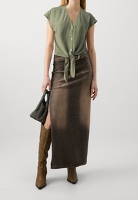 Grüne Kurzarmbluse mit einem Knoten in der Taille, kombiniert mit einem braunen, gradienten Jeans-Maxikleid und knielangen braunen High-Heels-Stiefeln.